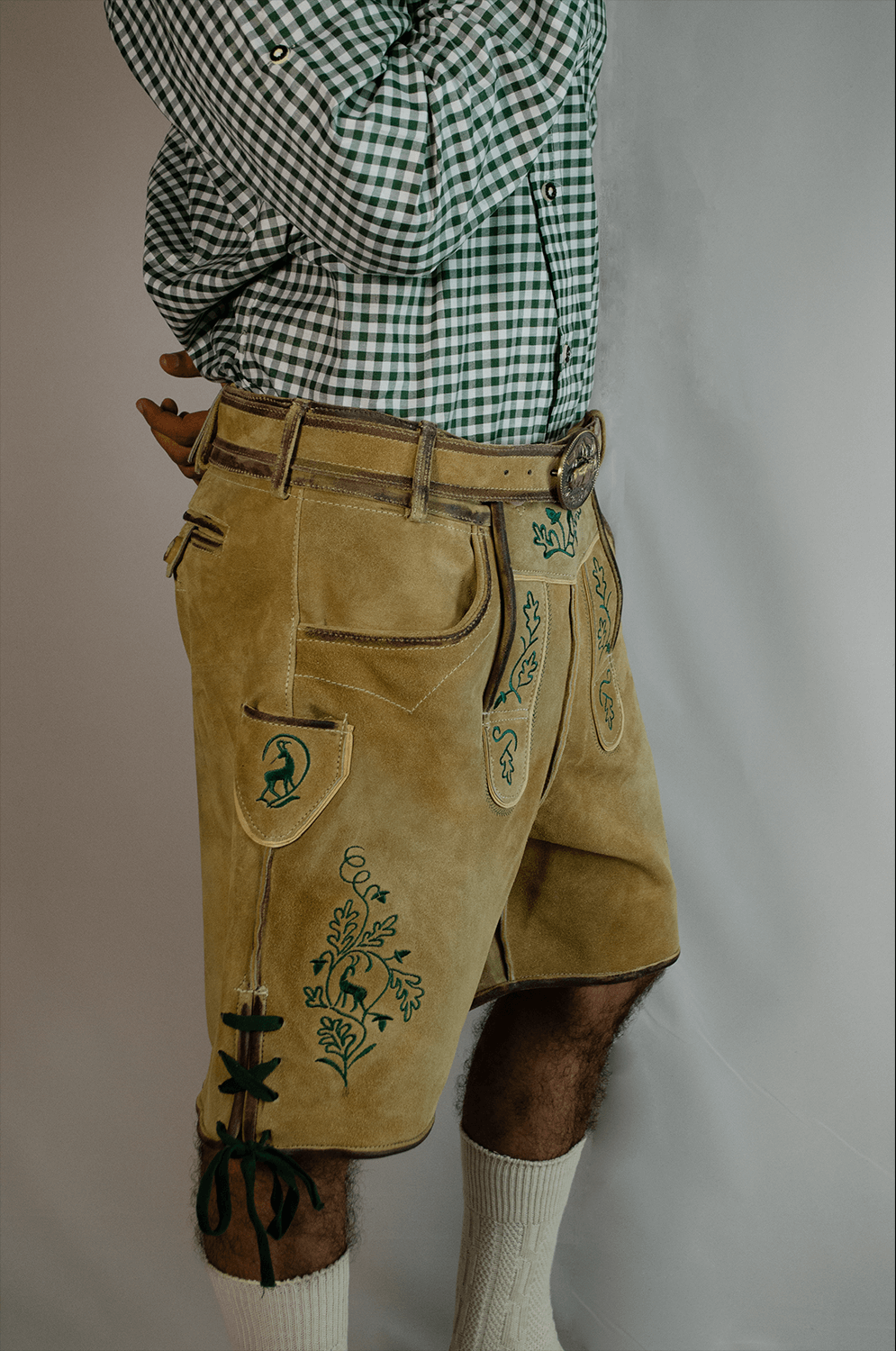Side view of a man wearing light gold handcrafted Lederhosen shorts with detailed green stag and vine embroidery, a matching leather belt, and a green checkered shirt. - Lederhosen For Sale