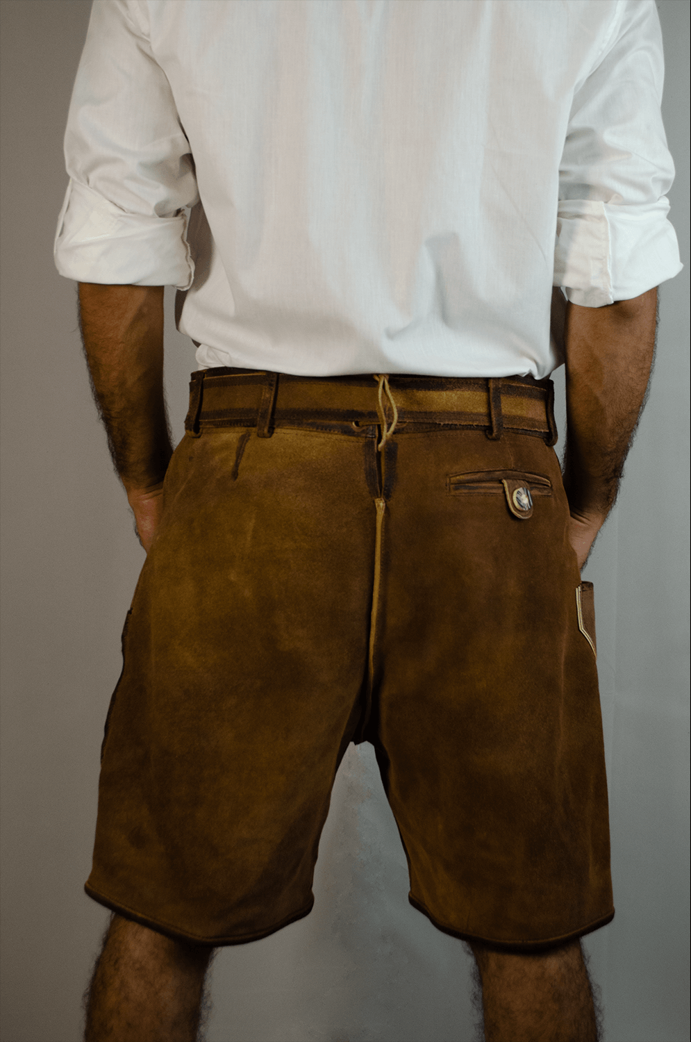 Back view of a man wearing vintage brown suede Lederhosen shorts with a matching belt and a white button-down shirt. - Lederhosen For Sale