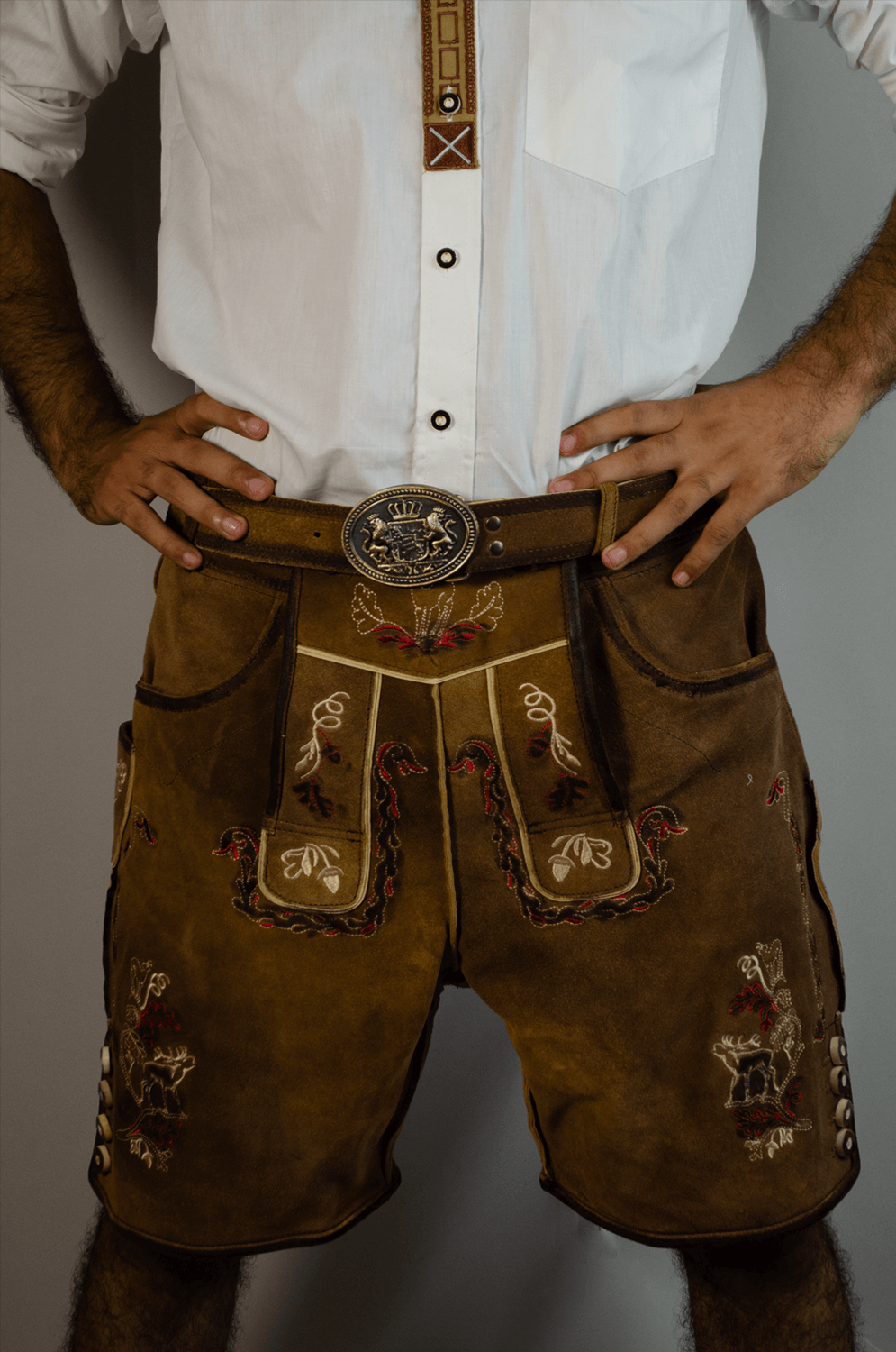 Front close-up of a man wearing vintage brown Lederhosen shorts with multi-colored embroidery, a matching leather belt, and a large ornate silver buckle. - Lederhosen For Sale